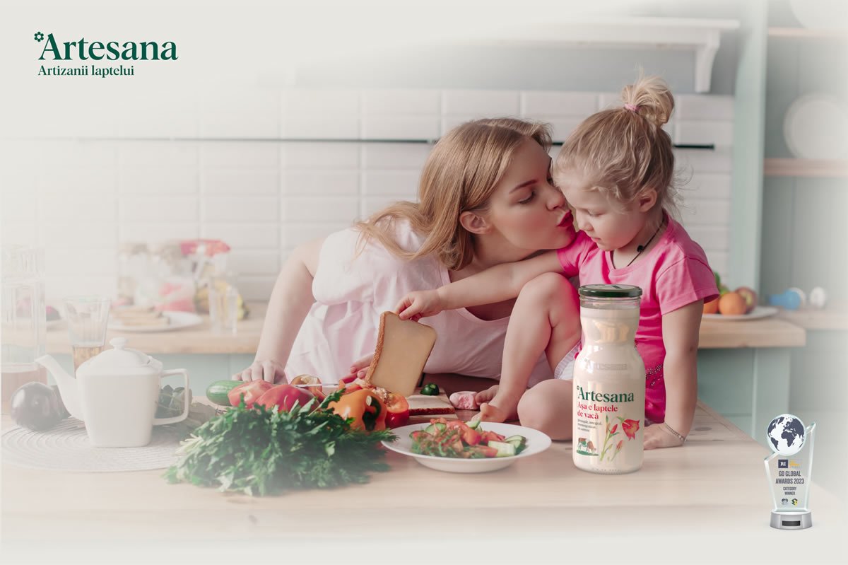 Mamă care își sărută fetița în bucătărie, lângă o sticlă de lapte Artesana din lapte de vacă, în timpul pregătirii micului dejun cu ingrediente proaspete.