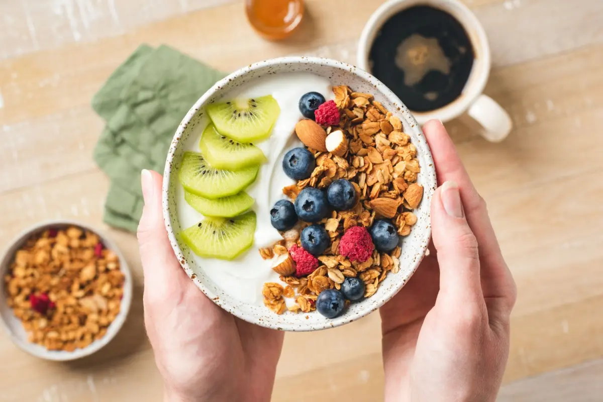 femeie ținând un bol cu iaurt, granola, fructe si nuci in maini