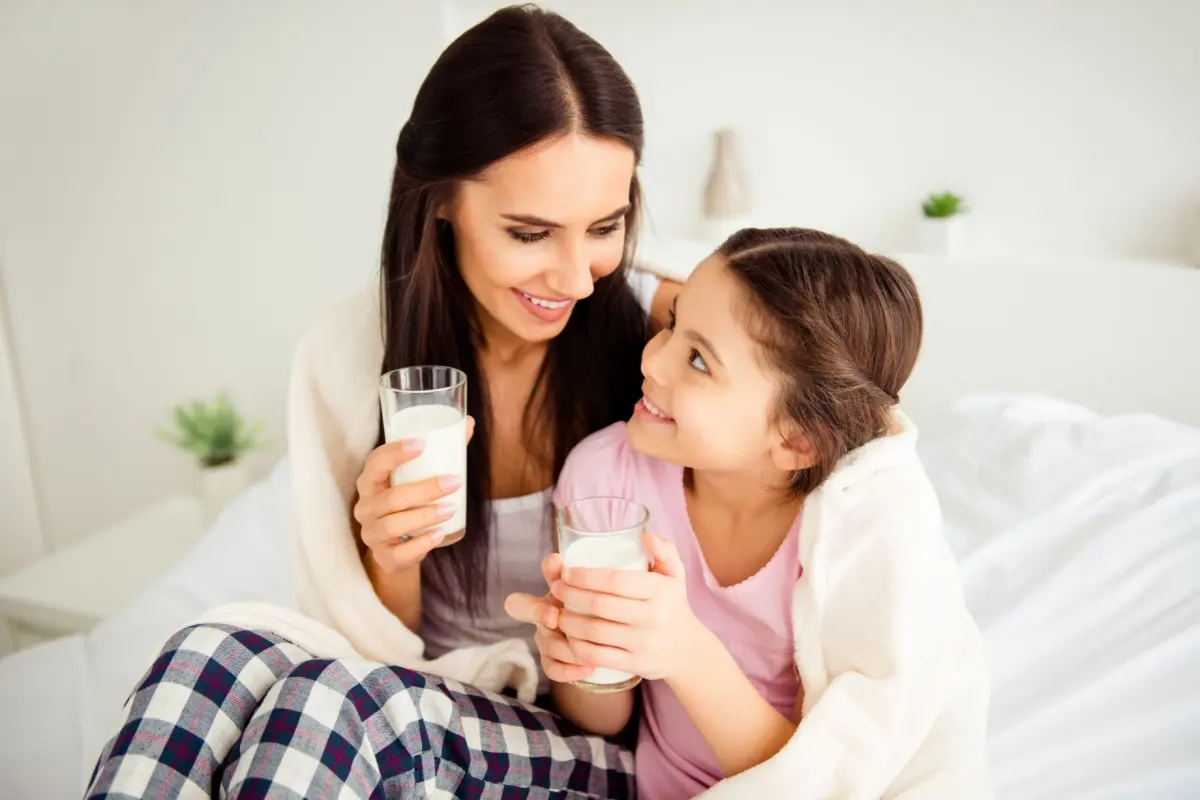 Mama si fiica stand pe canapea invelite in patura si band kefir din pahare.