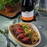 O portie de aripioare de pui caramelizate Fragedo cu o sticla de vin in fundal