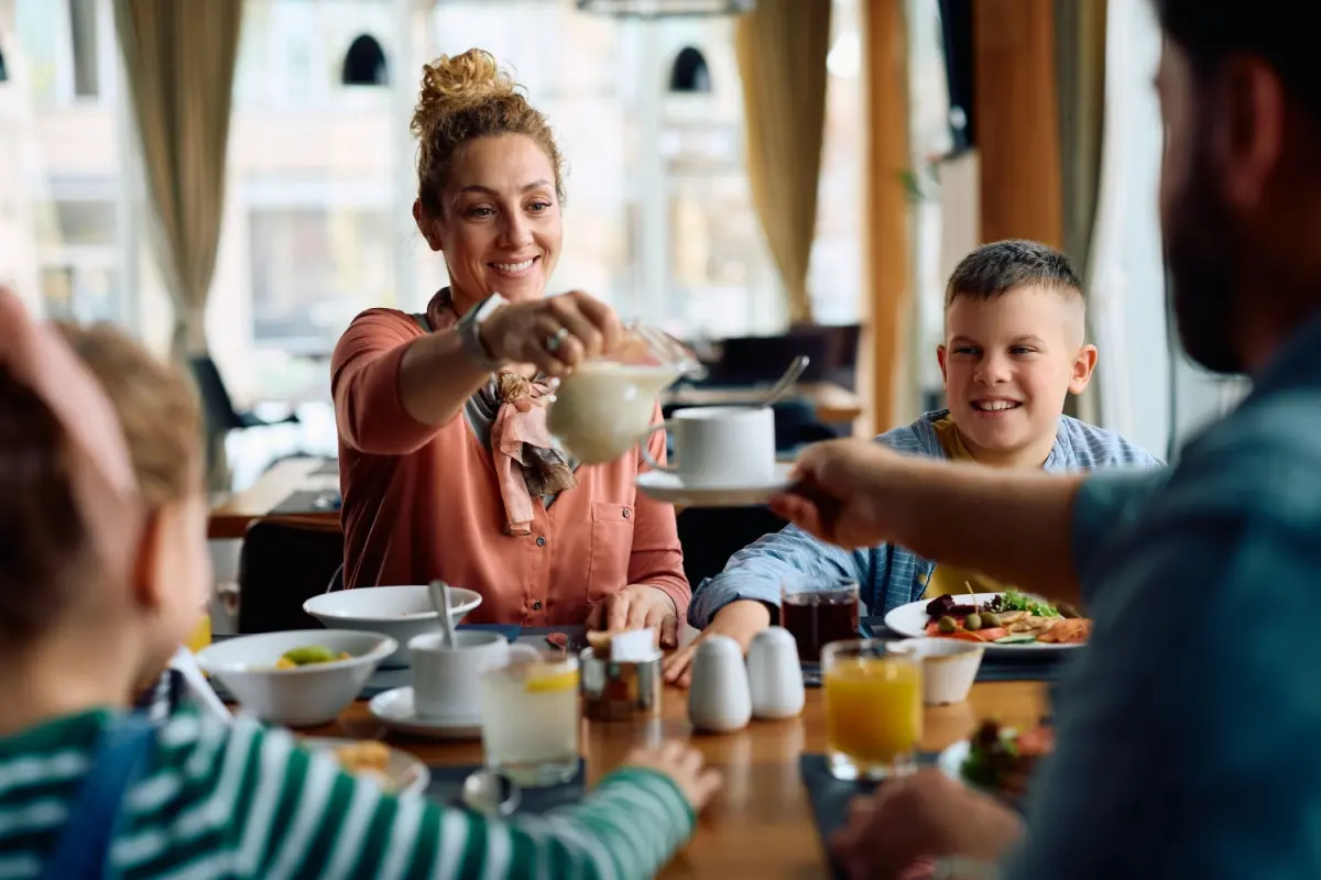 Familie de patru persoane servind mic-dejunul la restaurant. Femeia îi toarnă soțului lapte în cafea.