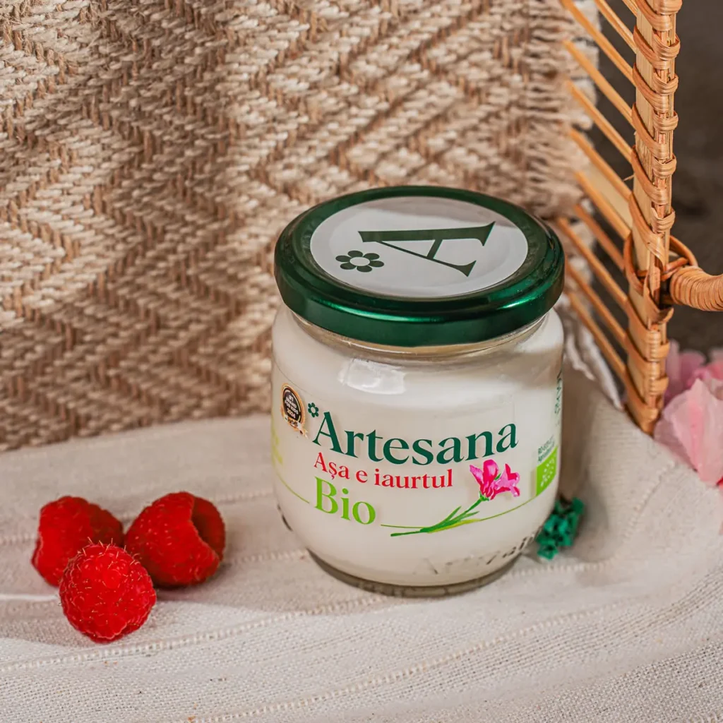 Un borcan de iaurt de vaca certificat ecologic Bio Artesana, la 160g, langa trei fructe de zmeura, cu un fundal de cos impletit in spate