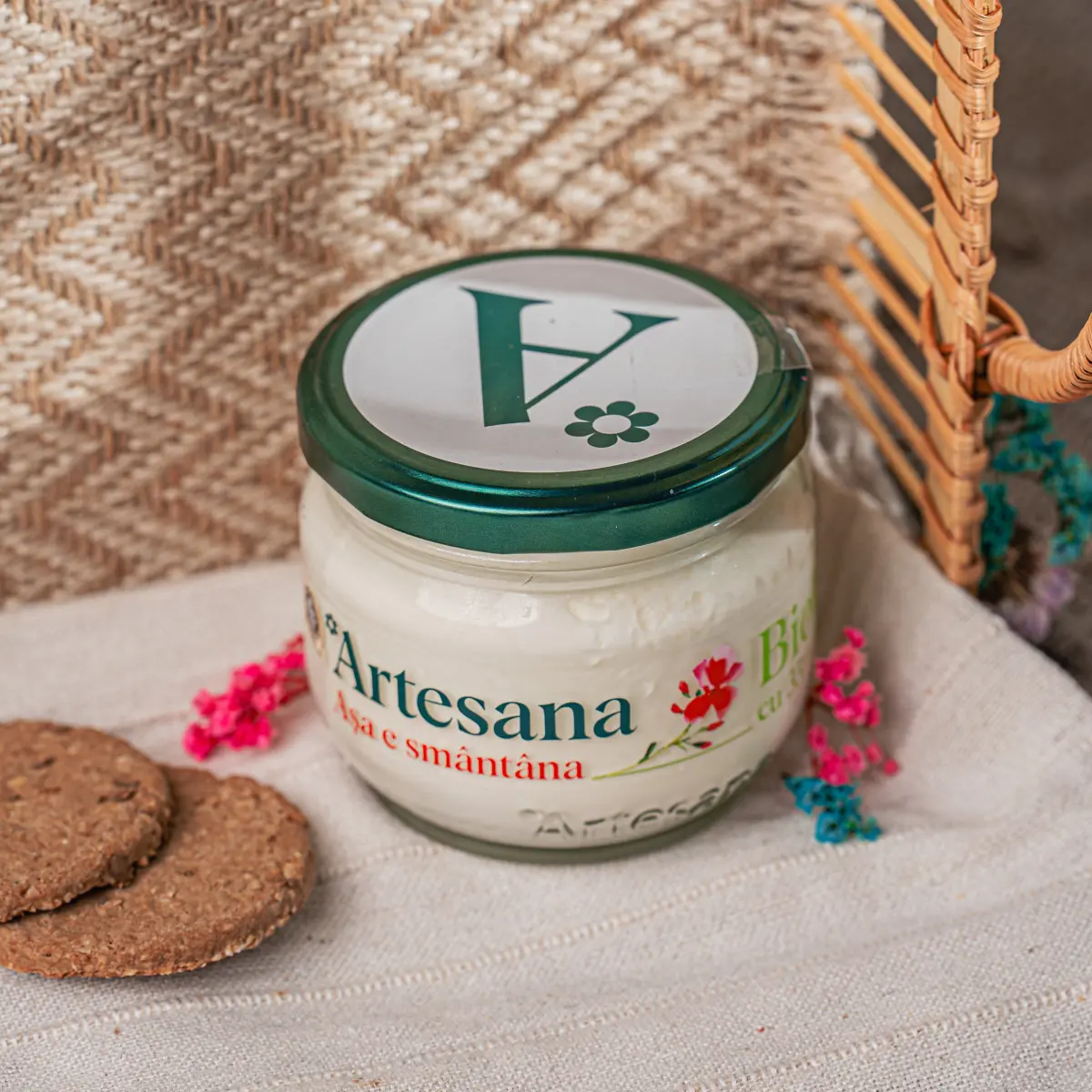 Un borcan de smantana Bio Artesana pe un fundal textil langa un biscuite si flori de camp colorate