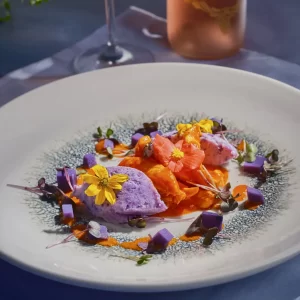 Preparat culinar gourmet cu piept de pui Fragedo în sos roze, quenelle de cartofi violet, cuburi de cartof mov și flori comestibile galbene și roz, servit elegant pe o farfurie albă decorată artistic.