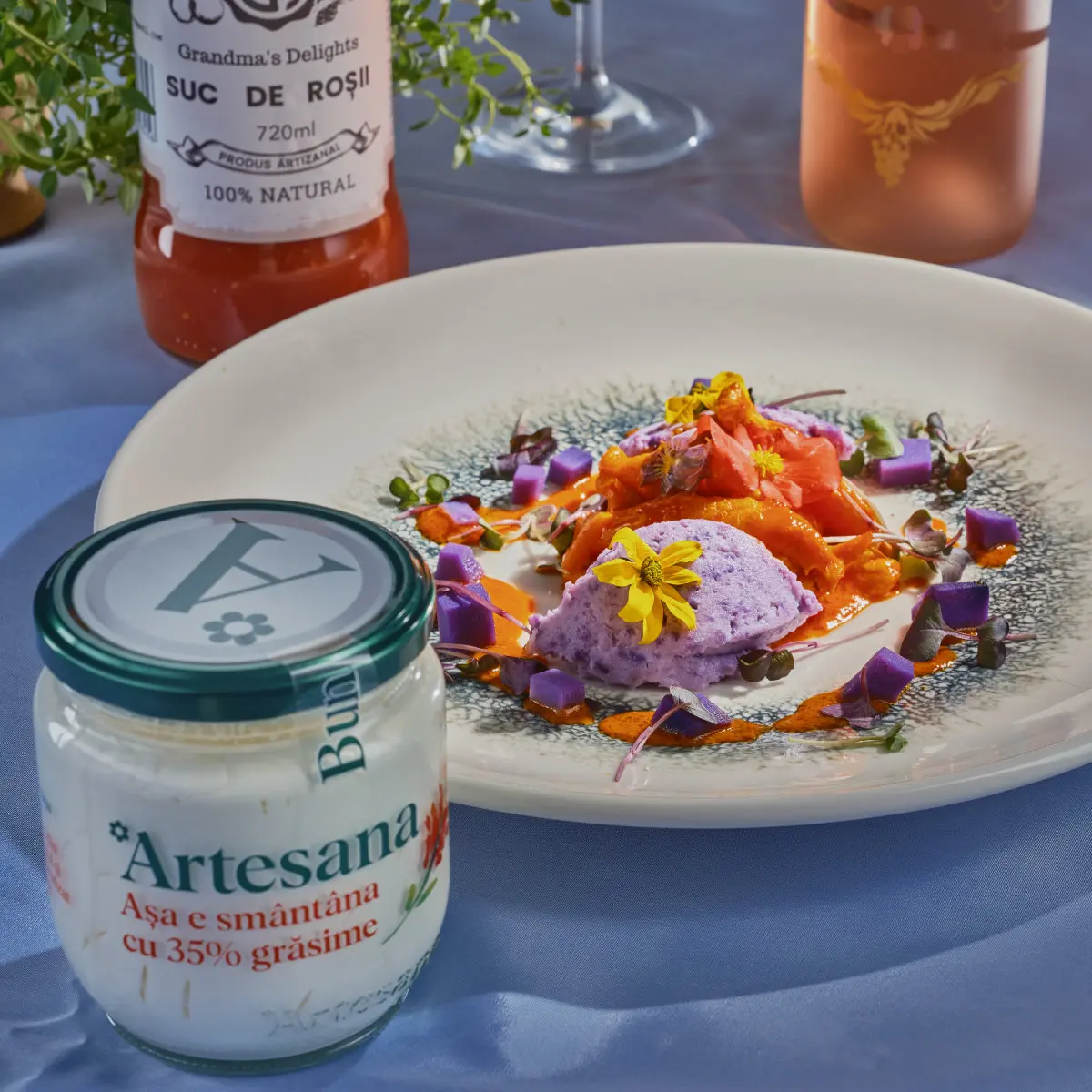 Borcan de smântână Artesana cu 35% grăsime alături de un preparat gourmet cu piept de pui Fragedo, sos roze, quenelle de cartofi violet și flori comestibile, pe fundal cu suc de roșii artizanal.