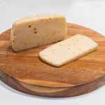 Brânză maturată de vacă cu peperoncino Artesana, așezată pe un platou rotund din lemn, prezentând textura fină și bucățelele de ardei iute din compoziție.