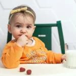 Bebeluș care stă la masă și mănâncă, purtând o bluză portocalie, cu un pahar de lapte și câteva zmeure