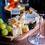 Un platou elegant cu brânzeturi maturate Artesana, tăiate în cuburi uniforme și aranjate apetisant pe un tocător din lemn, alături de struguri verzi și prune uscate. Două stegulețe cu logo-ul Artesana evidențiază originea artizanală a produselor. În prim-plan se observă piciorul unui pahar de vin roșu, sugerând o degustare gourmet într-o atmosferă caldă, festivă. Lumina blândă și fundalul decorativ creează un cadru premium, ideal pentru promovarea brânzeturilor maturate și a experiențelor rafinate oferite de Artesana.