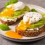 Tartine cu cremă de brânză, avocado feliat și ou poșat cu gălbenuș curgător, servite pe pâine integrală cu semințe.