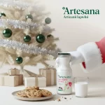 Sticlă de lapte Artesana „Așa e laptele de vacă”, ținută de Moș Crăciun, lângă un pahar cu lapte și fursecuri, într-un decor de Crăciun cu brad și cadouri.