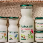 Gama de produse lactate bio Artesana – lapte integral cu caimac, sana bio, kefir bio, iaurt bio și smântână bio, ambalate în sticlă, prezentate pe fundal textil natural.