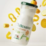 Sticlă de iaurt de băut Artesana „Așa e sănă” BIO, realizat din lapte ecologic, cu legume galbene ilustrate în jur – porumb, ardei și roșie galbenă, pe fundal deschis.