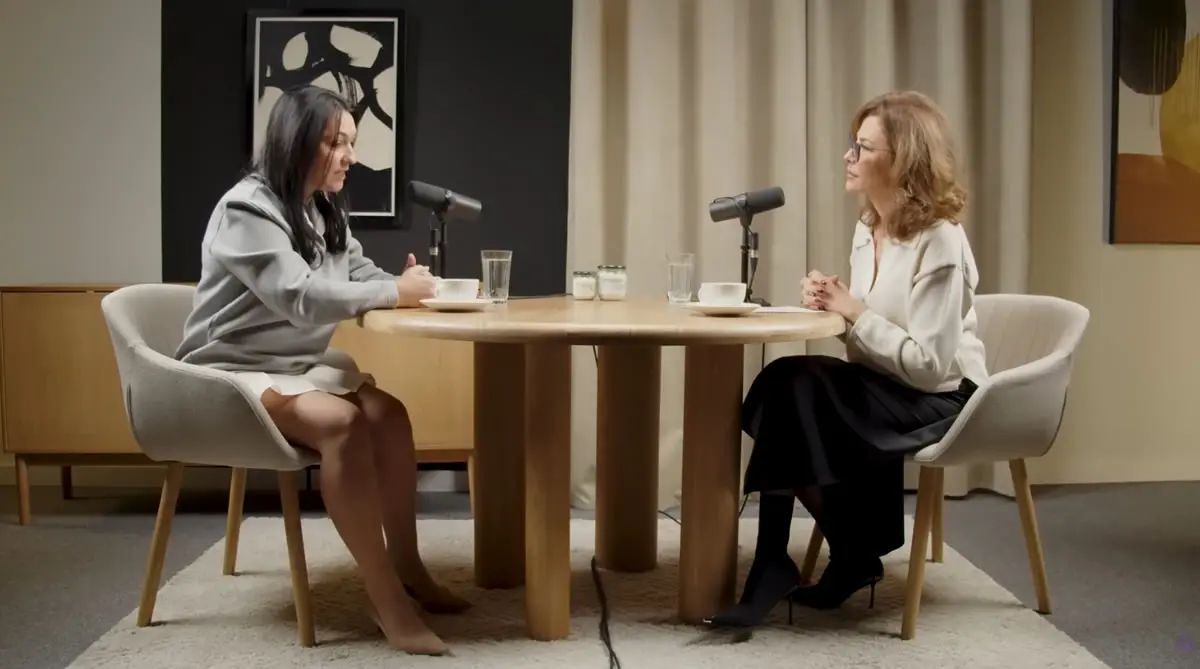 Alina Donici și Adina Alberts în timpul unui podcast, așezate față în față la o masă rotundă, cu microfoane profesionale și cești de cafea, într-un studio elegant.