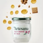 Borcan de iaurt natural de vacă Artesana, în ambalaj de sticlă cu capac verde, cu fulgi de porumb, granola și biscuiți plutind deasupra pe fundal deschis