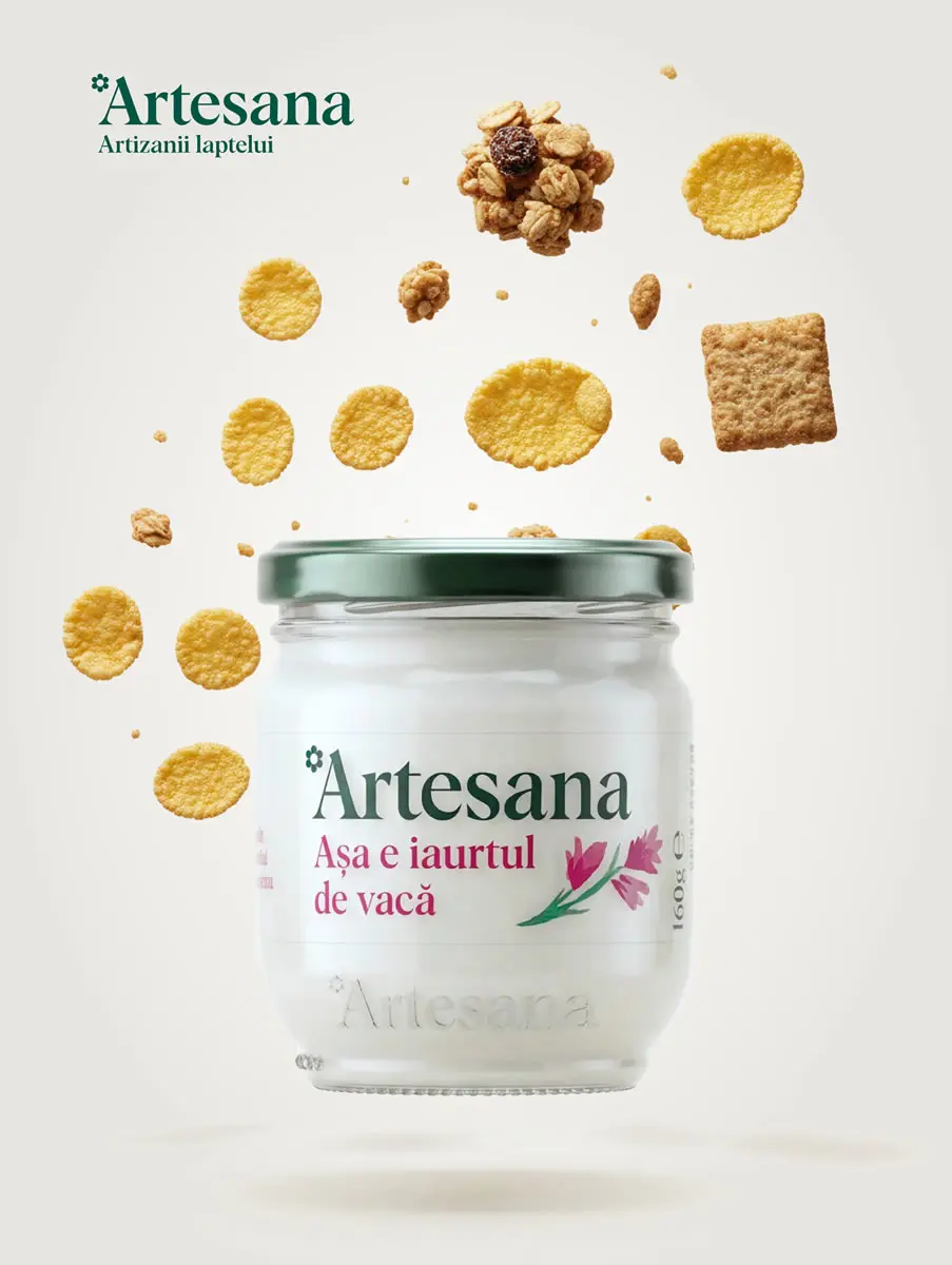 Borcan de iaurt natural de vacă Artesana, în ambalaj de sticlă cu capac verde, cu fulgi de porumb, granola și biscuiți plutind deasupra pe fundal deschis