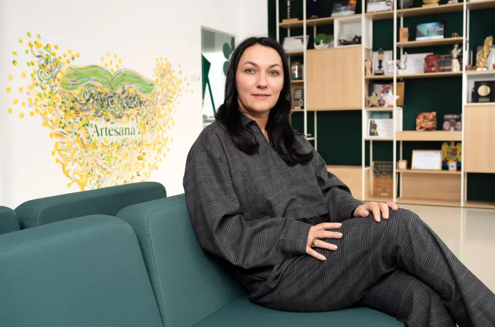 Alina Donici, cofondatoarea Artesana, fotografiată în sediul companiei, așezată pe o canapea verde, cu logo-ul Artesana ilustrat artistic pe perete și rafturi cu premii în fundal. Sursa foto: Forbes