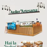 Stand Atelier Artesana amplasat în incinta Carrefour Băneasa, București, cu vitrină frigorifică din lemn și sticlă, logo „Artesana – Artizanii laptelui” și mesajul „Hai la Atelier!”. În prim-plan sunt expuse pe platouri din lemn produse lactate artizanale: brânzeturi maturate și proaspete, telemea, caș, unt și borcan cu specialitate Artesana. Pe stand apare mențiunea „integral, neomogenizat, pasteurizat la temperatură blândă, cu caimac”.