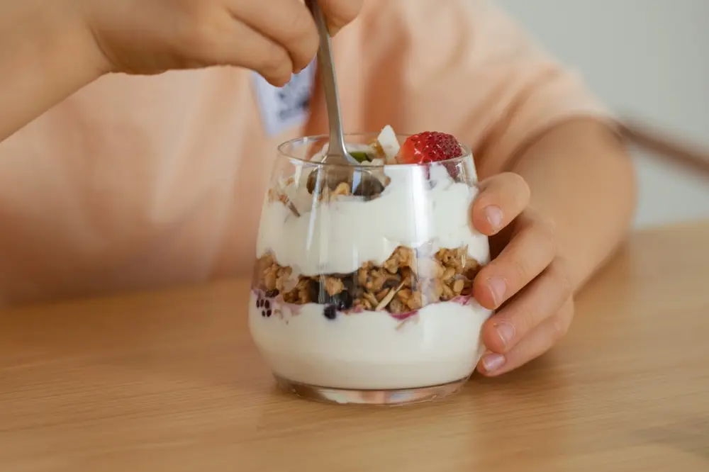 Copil mâncând iaurt cu granola și fructe proaspete, servit într-un pahar de sticlă, gustare sănătoasă pentru micul dejun sau pachețelul de școală.