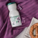 Sticlă de kefir de capră Artesana, din lapte integral și neomogenizat, 350 ml, așezată pe material mov texturat, lângă un covrig cu semințe.