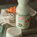 Sticlă de lapte de vacă Artesana, integral și neomogenizat, cu caimac, alături de un pahar cu lapte și un croissant, pe fundal textil în tonuri calde.