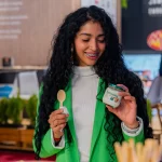 Femeie zâmbitoare degustând iaurt Artesana într-un stand expozițional, ținând în mână un borcan de iaurt din lapte de vacă și o linguriță, într-un cadru modern de tip târg alimentar.