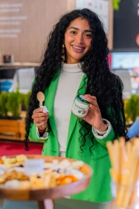 Femeie zâmbitoare degustând iaurt Artesana într-un stand expozițional, ținând în mână un borcan de iaurt din lapte de vacă și o linguriță, într-un cadru modern de tip târg alimentar.