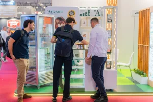 Vizitatori degustând produse lactate Artesana la stand expozițional, lângă vitrină frigorifică cu iaurturi și lapte, în cadrul unui târg internațional alimentar.