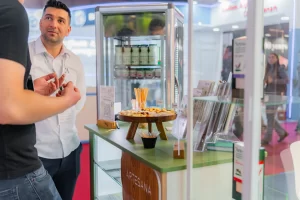 Degustare produse lactate Artesana la stand expozițional, cu mostre de iaurt și brânzeturi servite pe platou, vitrină frigorifică cu sticle de lapte și materiale de prezentare în cadrul unui târg alimentar.