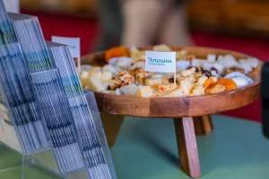 Platou de degustare cu brânzeturi și gustări Artesana, prezentat pe suport din lemn la stand expozițional, alături de pliante informative, în cadrul unui târg alimentar.
