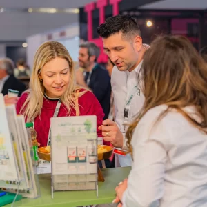 Vizitatori discutând și degustând produse lactate Artesana la stand expozițional, cu materiale informative și mostre de brânzeturi, în cadrul unui târg internațional alimentar.