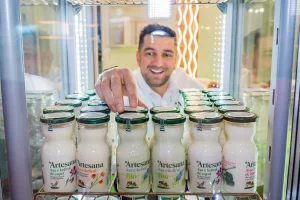 Reprezentant Artesana aranjând sticle de chefir și iaurt în vitrină frigorifică, evidențiind produse lactate artizanale Bio la stand expozițional din cadrul unui târg alimentar.