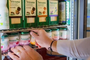 Mână aranjând borcane de iaurt Artesana cu vișine într-o vitrină frigorifică, alături de brânzeturi maturate artizanale, expuse la stand într-un târg alimentar.