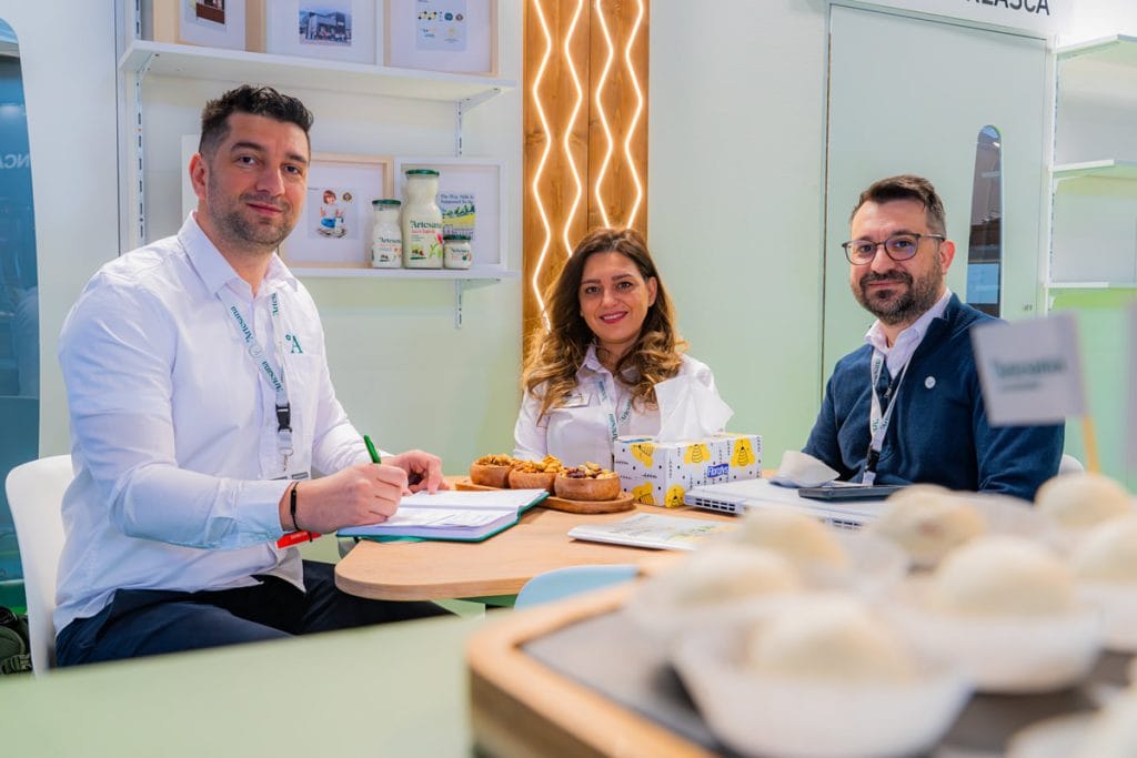 Echipa Artesana la târg alimentar, în discuție la masă, cu degustare de produse lactate artizanale și branding Milk Artisans vizibil în stand.