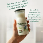 Mână ținând o sticlă de kefir Bio Artesana din lapte de vacă, produs artizanal, pasteurizat la temperatură blândă, ambalat în sticlă, brand Artesana - artizanii laptelui, mesaj despre produse făcute lent și cu grijă