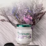 Iaurt de capră Artesana în borcan de sticlă, cu capac verde, decorat cu flori mov și roz, așezat pe material textil deschis la culoare, alături de o lingură neagră – produs artizanal natural din lapte de capră