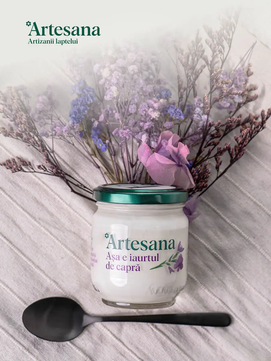 Iaurt de capră Artesana în borcan de sticlă, cu capac verde, decorat cu flori mov și roz, așezat pe material textil deschis la culoare, alături de o lingură neagră – produs artizanal natural din lapte de capră