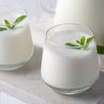 Două pahare de chefir cremos din lapte de vacă, servite reci și decorate cu frunze proaspete de mentă, alături de o sticlă cu chefir pe fundal, produs lactat fermentat natural.