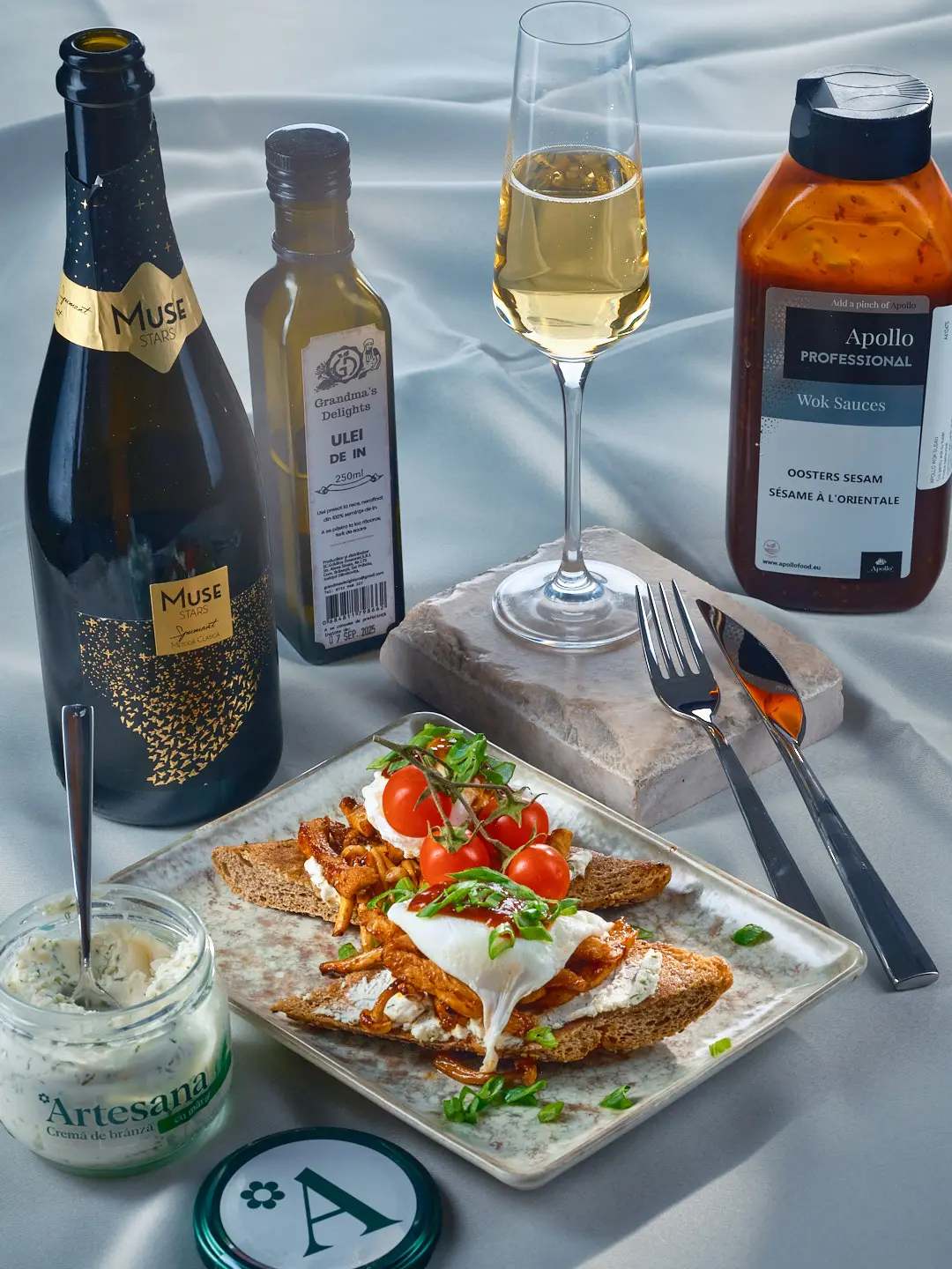 Bruschete gourmet cu cremă de brânză Artesana cu mărar, piept de pui fraged în stil oriental cu ciuperci shimeji, ou poșat și roșii cherry, servite alături de vin spumant și ingrediente premium.