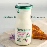 Sticlă de sana de capră Artesana din lapte integral și neomogenizat, așezată pe o pânză naturală lângă un croissant, cu mesajele „Hidratează-te” și „Hrănește-te”, promovând produse lactate artizanale naturale.