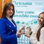 Două tinere prezintă produse lactate Artesana artizanale la standul brandului în cadrul târgului TuttoFood Milano 2026, ținând în mâini iaurt și lapte bio Artesana, pe fundalul unui panou cu mesajul „Il latte come dovrebbe essere”.