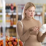 Femeie într-un supermarket care citește eticheta unui pachet de iaurt înainte de cumpărare, verificând ingredientele și informațiile nutriționale.
