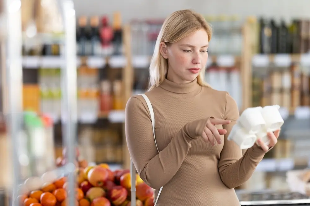 Femeie într-un supermarket care citește eticheta unui pachet de iaurt înainte de cumpărare, verificând ingredientele și informațiile nutriționale.