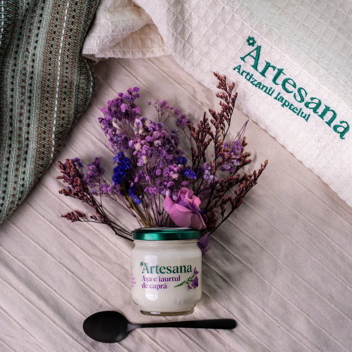 Borcan de iaurt de capră Artesana 160 g din lapte integral, minim procesat, prezentat pe un fundal textil alături de flori uscate și o linguriță, într-un decor rustic.