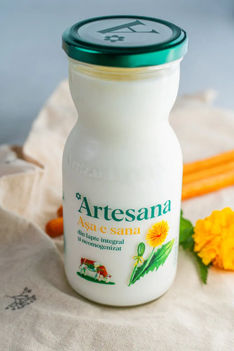 Sticlă de Sana Artesana 350 ml, obținută din lapte integral românesc, neomogenizat, fermentată natural și ambalată în sticlă, prezentată alături de flori și elemente naturale.