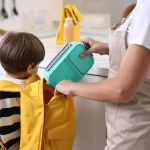 Părinte pregătește pachetul pentru școală și pune o caserolă reutilizabilă în ghiozdanul unui copil, într-o bucătărie luminoasă, ilustrând obiceiuri sănătoase și organizarea mesei pentru școală.