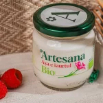 Borcan cu iaurt Bio Artesana din lapte ecologic, în ambalaj de sticlă, așezat pe un șervet textil alături de zmeură proaspătă.