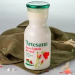Sticlă de lapte integral de vacă Artesana, neomogenizat și cu caimac, așezată pe un fundal textil verde, decorat cu flori.