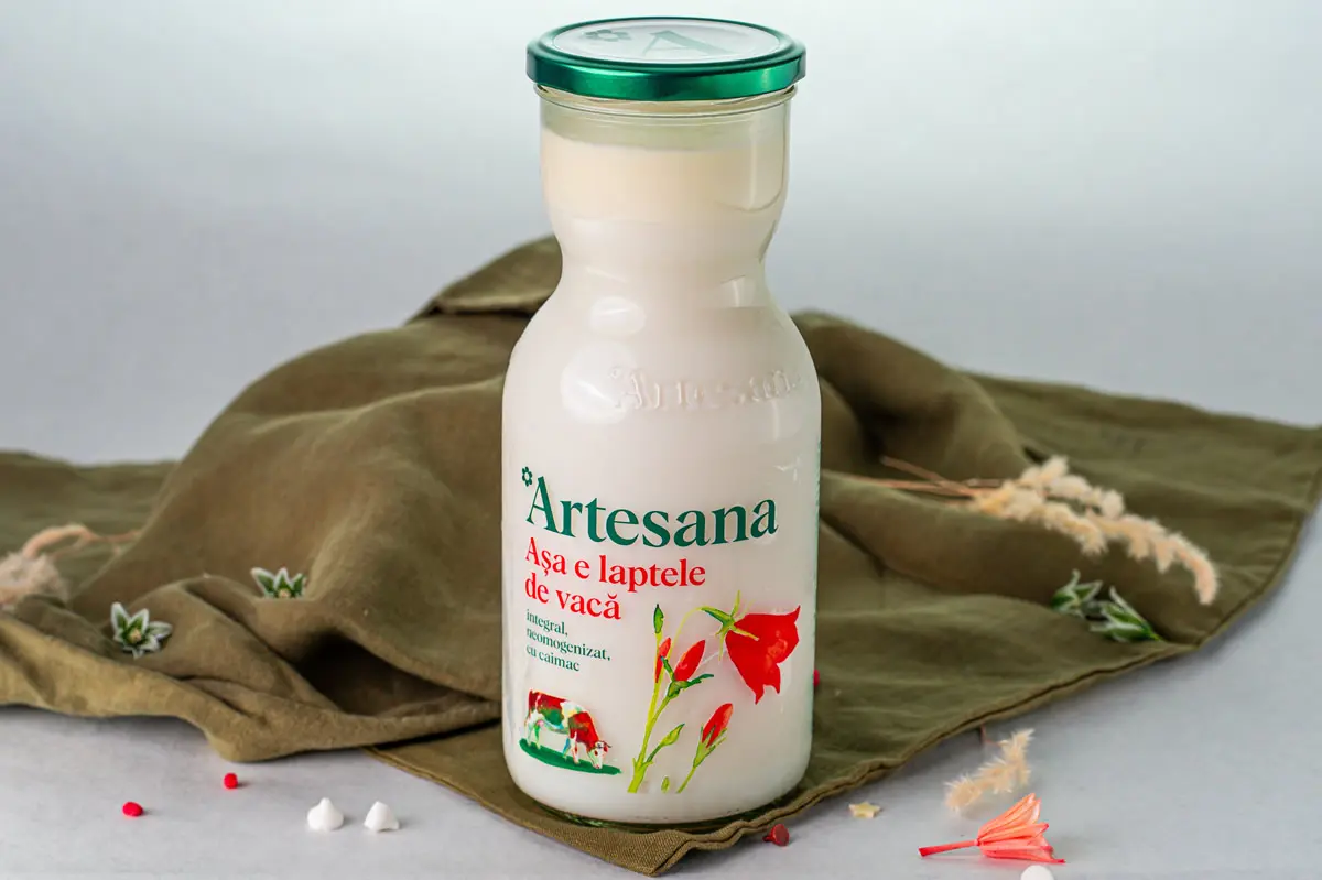 Sticlă de lapte integral de vacă Artesana, neomogenizat și cu caimac, așezată pe un fundal textil verde, decorat cu flori.
