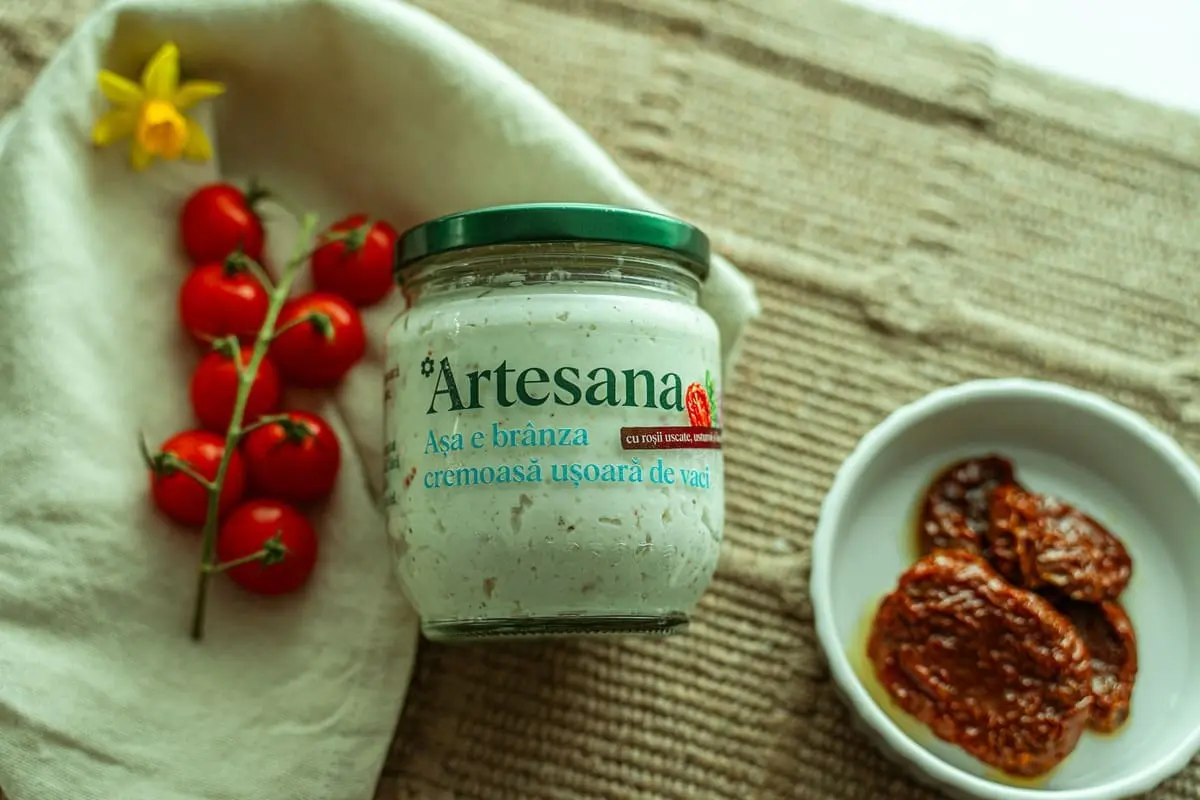 Brânză cremoasă ușoară de vaci Artesana cu roșii uscate, usturoi și busuioc, în borcan de sticlă, alături de roșii cherry și un bol cu roșii uscate.
