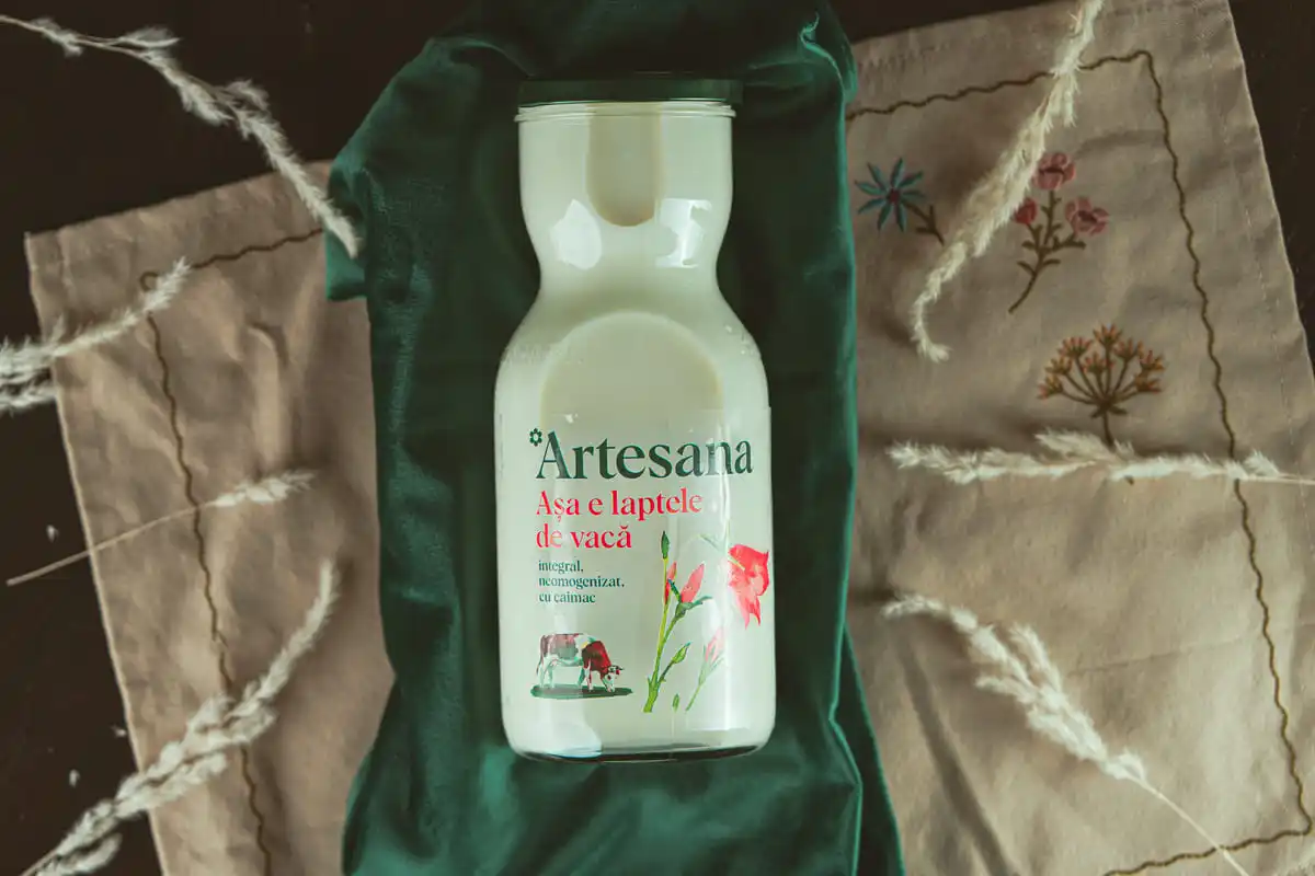Sticlă de lapte de vacă Artesana, așezată pe un material verde și un șervet textil decorat cu broderii florale și spice uscate.